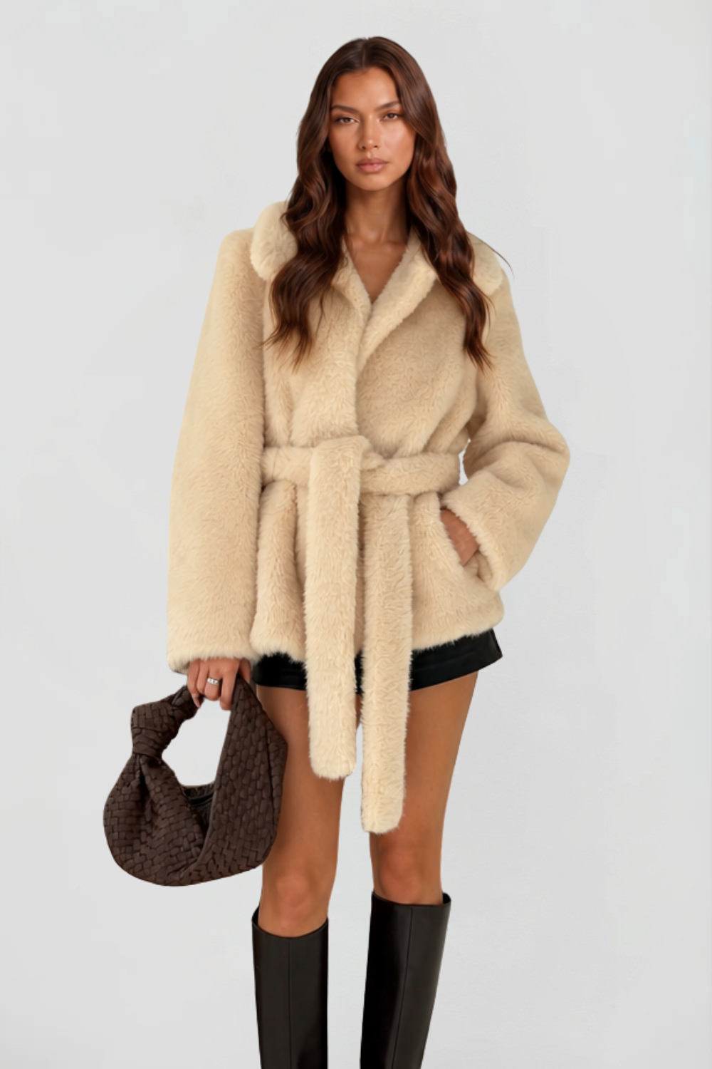 Woman wearing cream shearling belted short coat with notch lapel holding a woven brown leather bag