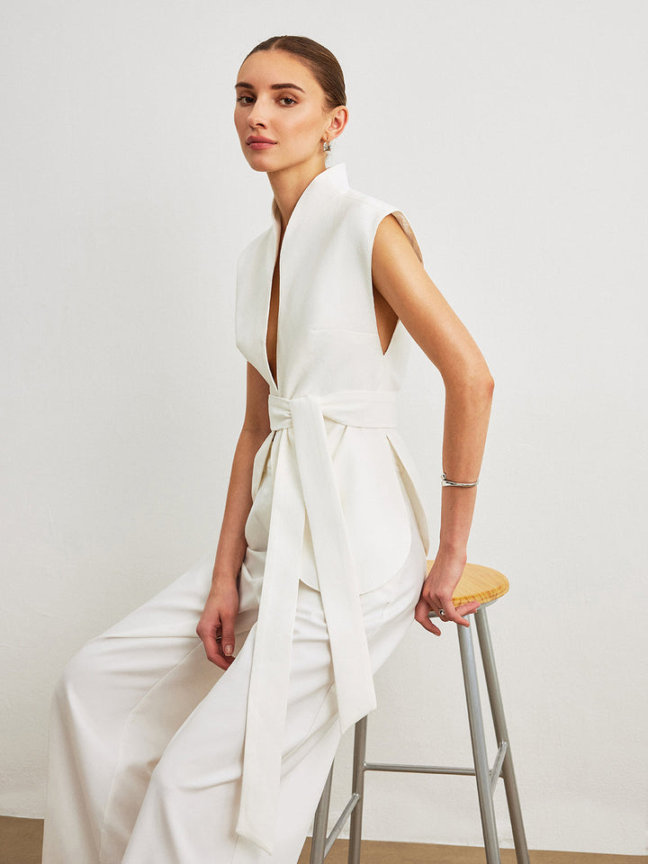 Model sits on a stool in white sleeveless wrap top with self-tie waist belt