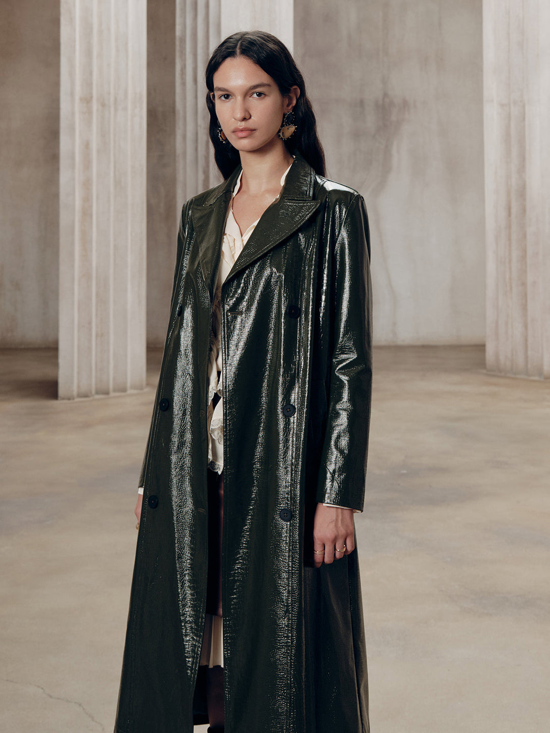 Army green glossy PU leather trench coat, worn open full length against a stone colonnade
