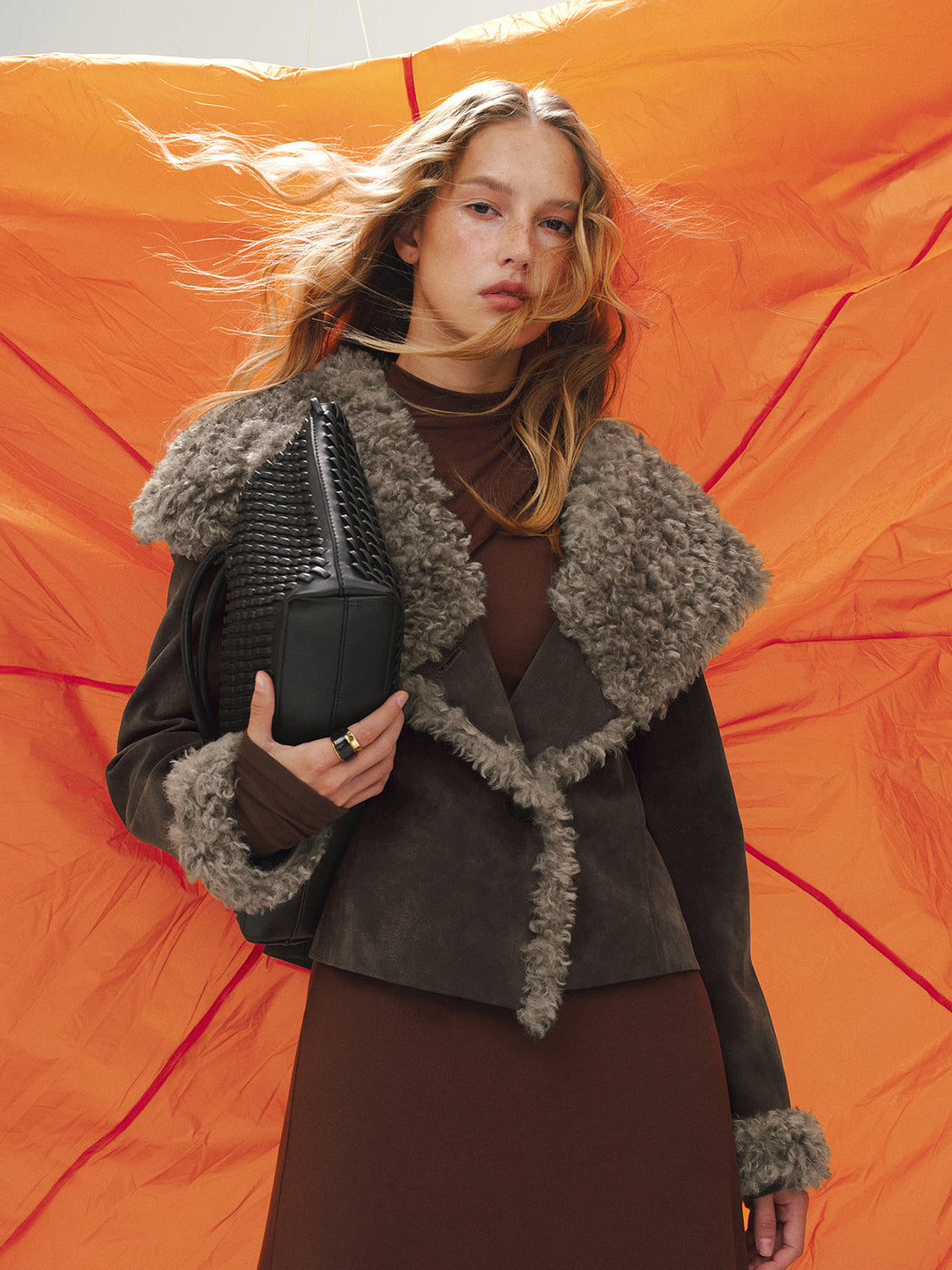Model in shearling collar suede jacket against orange backdrop, holding a black woven clutch