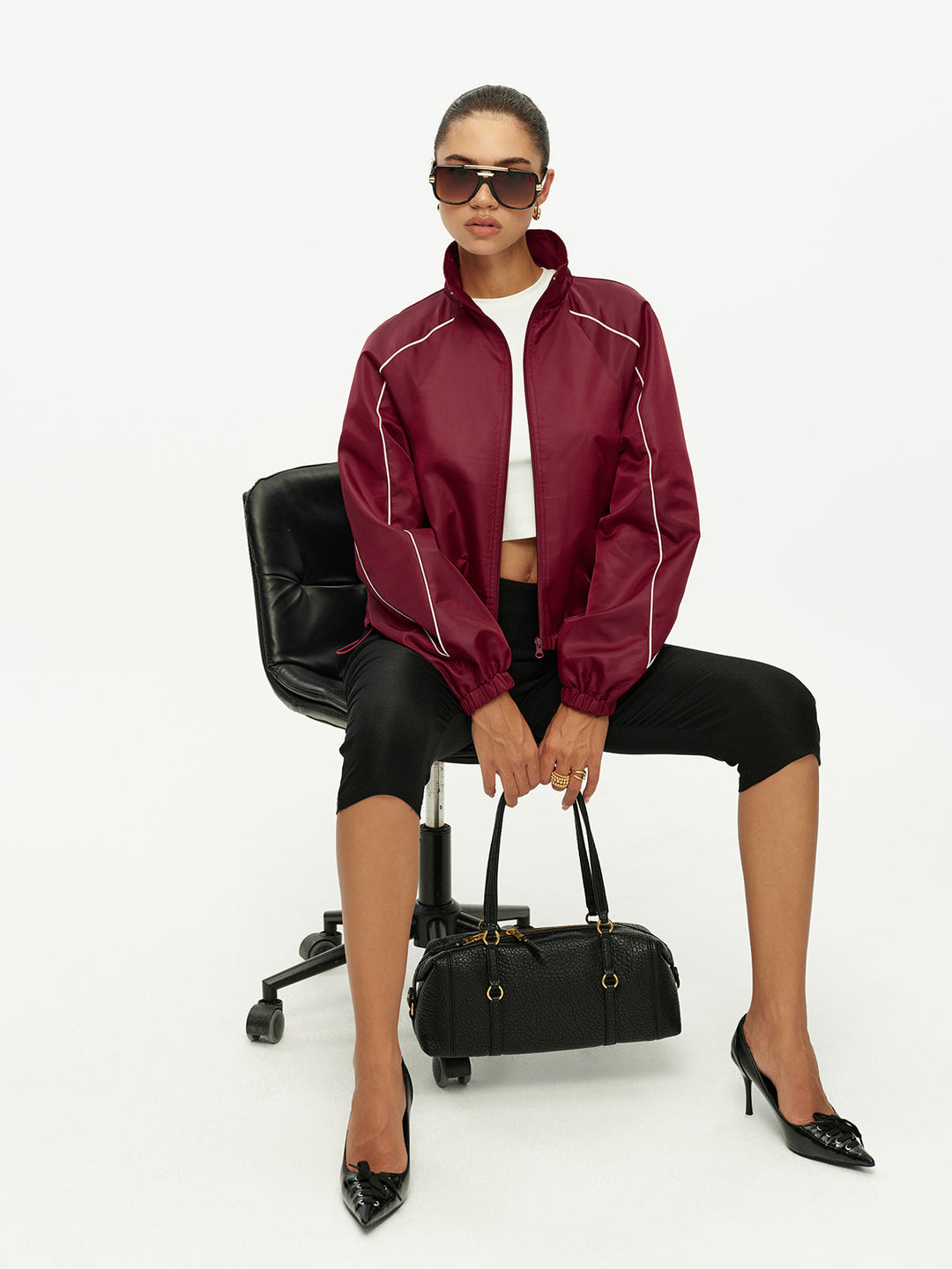 Model seated wearing burgundy piped track jacket with white crop top and black capris