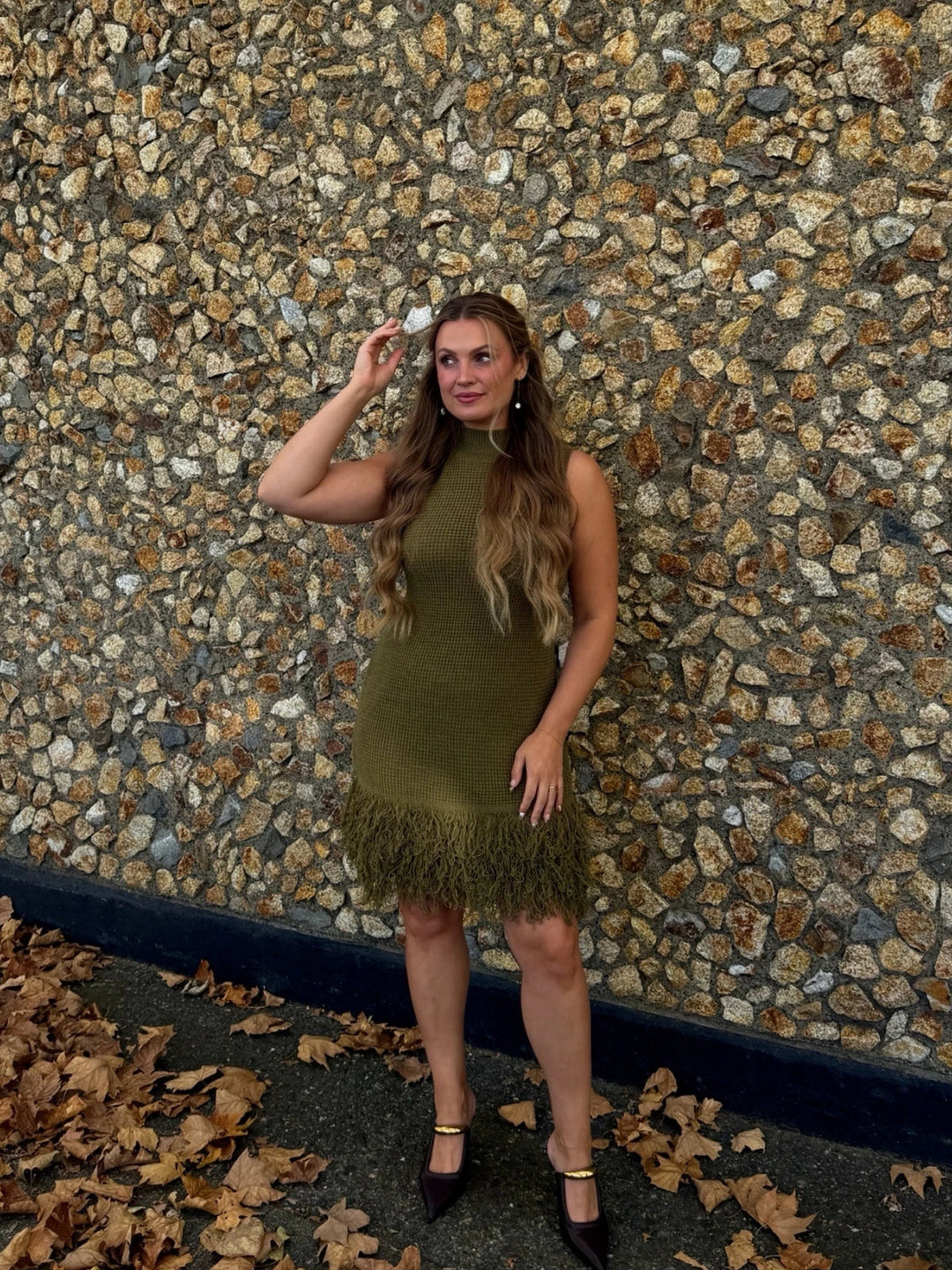 Model posing outdoors in an olive green mock neck knit dress with feathered fringe hem